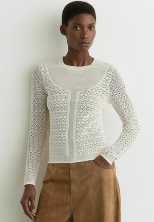 Mujer con camiseta blanca de manga larga con patrón de encaje y pantalones de ante marrones, de pie contra una pared lisa y clara con una mano en el bolsillo.