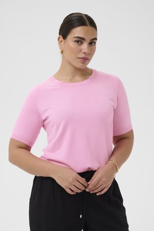 Femme aux cheveux foncés tirés en arrière, portant un haut en tricot rose à manches courtes et un pantalon noir, tenant ses mains près de sa taille.