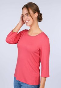 Korallenfarbenes Langarmshirt mit Rundhalsausschnitt und dekorativen Nähten. Hergestellt aus einem glatten Material, kombiniert mit blauen Jeans.