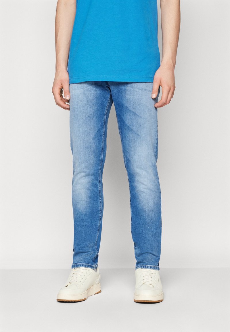 Personne portant un jean slim bleu clair, des baskets blanches et un t-shirt bleu, debout contre un fond blanc uni.