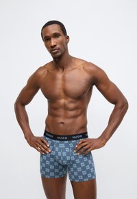 Calções boxer com padrão geométrico em azul e cinzento, com uma cintura elástica preta exibindo a marca "HUGO" em letras brancas.