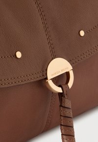 Καφέ δερμάτινη γυναικεία τσάντα με κυκλικό χρυσό μεταλλικό διακοσμητικό στοιχείο και υφή στην επιφάνεια, διακοσμημένη με διακριτικά χρυσά καρφιά και ραμμένες ραφές.