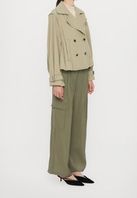 Veste courte vert olive à double boutonnage avec de gros boutons noirs, associée à un pantalon olive à jambes larges et des escarpins noirs à bout pointu.