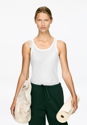 Weißes Baumwoll-Tanktop mit rundem Halsausschnitt, kombiniert mit dunkelgrünen Jogginghosen mit Kordelzug. Hält eine beige Tasche und eine gerollte Yogamatte.
