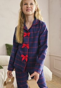 Námořnické modré kostkované pyžamo s červenými akcenty, obsahující knoflíky zdobené červenými mašličkami. Měkká tkanina a dlouhé rukávy.