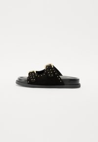 Sandalo in suede nero con doppie cinghie a fibbia e borchie dorate, dotato di una suola piatta nera. La vista laterale mette in risalto linee pulite e un design minimale.