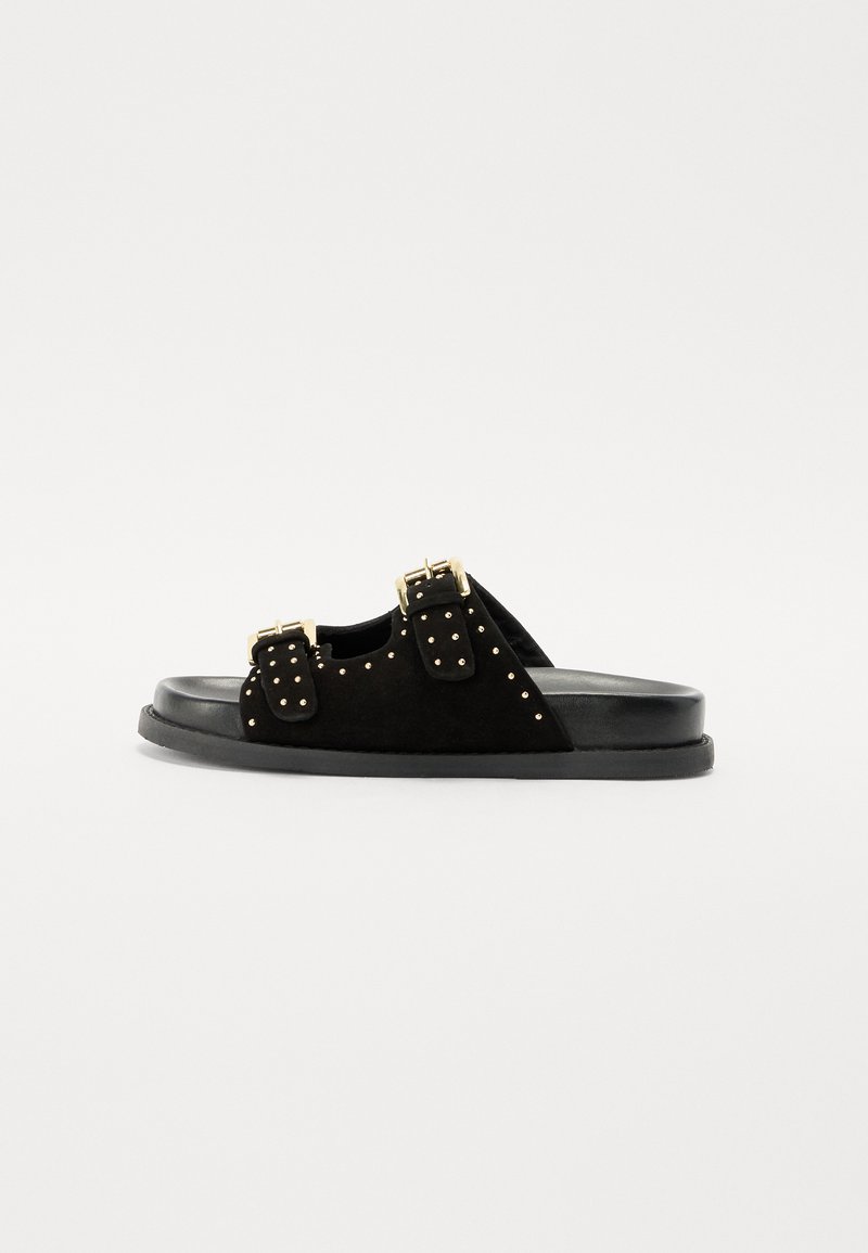 Sandalo in suede nero con doppie cinghie a fibbia e borchie dorate, dotato di una suola piatta nera. La vista laterale mette in risalto linee pulite e un design minimale.