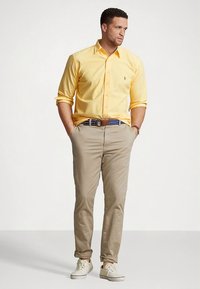 Κίτρινο πουκάμισο με κουμπιά, μπεζ chino παντελόνι, καφέ ζώνη και λευκά αθλητικά παπούτσια. Το μοντέλο στέκεται με τα χέρια στις τσέπες μπροστά σε ουδέτερο φόντο.