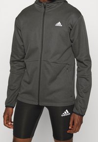 Chaqueta deportiva de color gris oscuro con cremallera, hecha de una tela texturizada, que cuenta con capucha y el logo blanco de Adidas en el pecho. La tela tiene un ligero brillo.