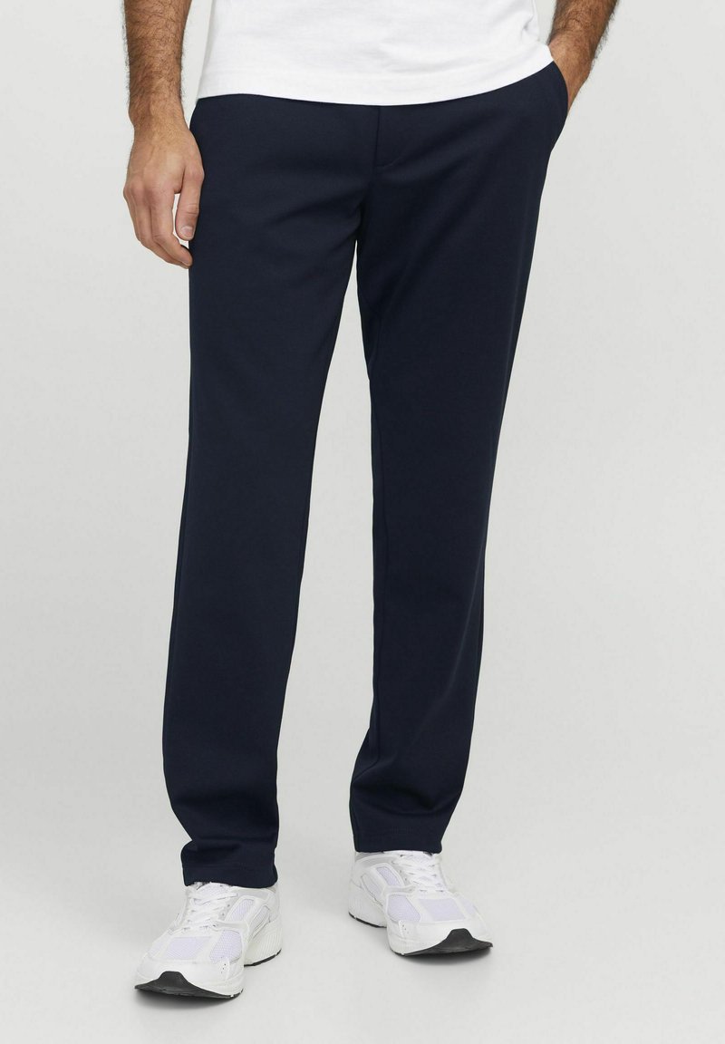Pantalons de survêtement bleu marine en tissu doux, présentant un design à jambe droite et des poches latérales. Associés à des chaussures de sport blanches.