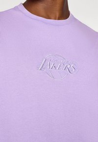 T-shirt lavande en coton avec un col rond. Présente un logo "Los Angeles Lakers" brodé en blanc accompagné d'un graphique de basket-ball.