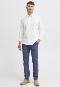 Chemise blanche à col, pantalon bleu et bottines beiges. La chemise a des manches longues ; le pantalon est ajusté et a une texture lisse.