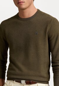 Olivgrön bomullströja i stickad kvalitet, med ribbad rund halsringning och långa ärmar, prydd med en liten broderad logotyp på bröstet.