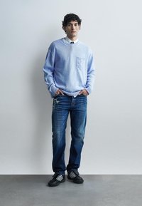 Jeune homme debout les mains dans les poches, portant un pull bleu clair, une chemise blanche avec une cravate noire, un jean déchiré et des baskets noires.