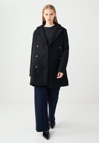 Schwarzer doppelt knöpfbarer Mantel aus einem Wollgemisch mit großen Knöpfen, Schalkragen und weiten Ärmeln. Getragen über einem dunklen Oberteil und weitem Jeans.