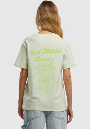 Mündroheline ülemõõdus T-särk, millel on graafika külma matcha latte'ist, kollaste aktsentidega ja tekst seljal. Lühikesed varrukad, puuvillane materjal.