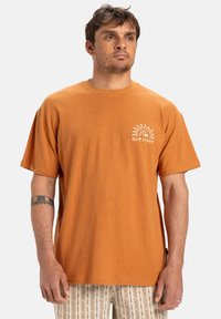 Krátký rukáv oranžového trička z bavlny, s bílou grafikou slunce a textem "Quiksilver" na levé části hrudi. Pohodlný střih.