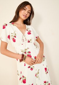 Haljina s cvjetnim uzorkom u bijeloj boji s ružičastim ružama i zelenim listovima. Odlikuje se V-izrezom, kratkim rukavima i detaljem za vezanje sprijeda. Glatki materijal.