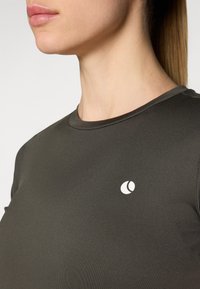 Tmavo zelené priliehavé tričko s okrúhlym výstrihom a malým bielym logom na ľavej strane. Hladká, ľahká tkanina.