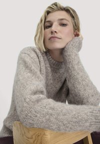 Hellbrauner Wollpullover mit rundem Halsausschnitt, gerippten Bündchen und einer weichen, strukturierten Oberfläche. Vor einem neutralen Hintergrund getragen, in lässigem Stil.