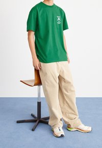 Camiseta de algodón verde con un pequeño gráfico en el pecho, combinada con pantalones de pana beige y zapatillas deportivas multicolores, sentado en una silla.