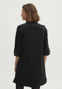 Sort skjortekjole lavet af blødt stof, med krave, opslåede ærmer og en buet kant. Rent, minimalistisk design uden synlige mønstre.