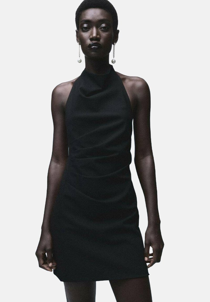 Vestido mini negro de tirantes con un tejido suave, con detalles fruncidos en el costado y una silueta ajustada. Presenta pendientes de perlas.