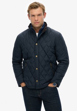 Navy quilted jacket with a stand collar, gold snap buttons, two side pockets, and contrasting brown detailing. Worn over denim.