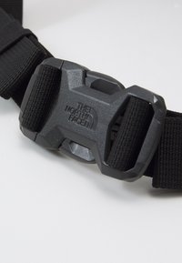 Juodas nailono diržas su matinio pilko plastiko užsegimu, ant kurio iškilmingai išgraviruotas logotipas „THE NORTH FACE“. Užsegimo dizainas apima ergonomines kraštines.