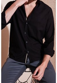 Mann trägt ein schwarzes Hemd mit aufgekrempelten Ärmeln und graue Hose, sitzt auf einem Drahtgestellstuhl vor einer strukturierten Wand.