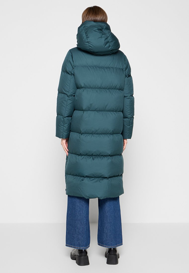 Langer, voluminöser türkisfarbener Mantel mit Kapuze, horizontal gestepptem Design, mittellanger Schnitt und getragen über blauen weiten Jeans.