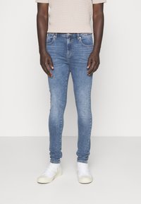 Jeans de mezclilla azul ajustados con un lavado desgastado, cinco bolsillos y trabillas para cinturón. Combinados con zapatillas blancas de caña baja y una camiseta de punto beige claro.