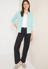 Cardigan bleu clair duveteux porté sur un haut blanc, associé à un pantalon large noir et des baskets beige avec des accents gris. Design simple et texture douce.