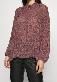 Fließende Bluse mit langen Ärmeln, die ein strukturiertes, gewelltes Muster in Rot- und Violetttönen aufweist. Mit rüschigem Ausschnitt und Schlüssellochdetail.