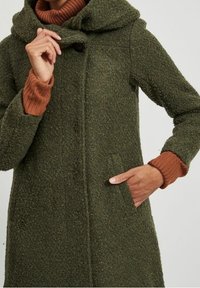 Cappotto in lana verde oliva con tessuto strutturato, cappuccio oversize, chiusura a bottoni e tasche laterali. Il modello indossa un maglione a costine color ruggine sotto.
