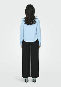 Blusa azzurra a maniche lunghe con orlo curvo, abbinata a pantaloni neri a gamba larga e scarpe con tacco teal, vista da dietro.