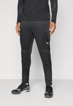 Pantalons de sport noirs en tissu extensible, dotés de détails rembourrés aux genoux et d'un petit logo blanc, associés à des chaussures de training noires.