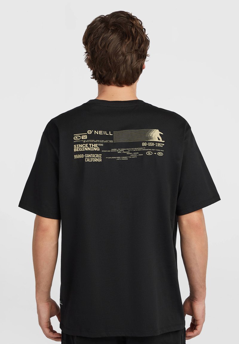 Άνδρας με μαύρο T-shirt που έχει μπεζ γραφικό θέμα surfing και κείμενο στο πάνω μέρος της πλάτης, στέκεται μπροστά σε απλό φωτεινό φόντο.