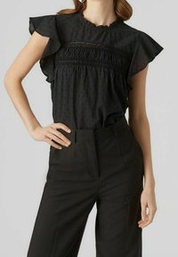Sort bluse med korte flæsekorte ærmer, struktureret stof og blonder detalje ved brystet. Kombineret med højtaljede sorte bukser.