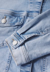 Giacca in denim blu chiaro con colletto, bottoni e cuciture visibili, caratterizzata da una texture leggermente consumata.
