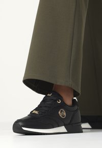 Černé tenisky s texturovaným povrchem, zlatými akcenty a bílou podrážkou. Nosené s kalhotami v olivově zelené barvě s širokými nohavicemi.