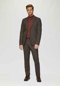 Bruin geruit pak met een getailleerde blazer en bijpassende broek, gecombineerd met een bordeauxrode turtleneck. Leren laarzen maken de outfit compleet.