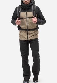 Męska kurtka w dwóch kolorach z czarną górą i brązowym dołem, z zapięciem na zamek i regulowanym paskiem. W połączeniu z czarnymi spodniami i trampkami.