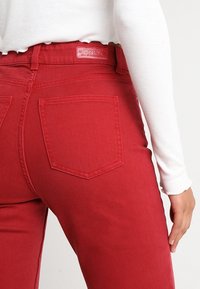 Jeans en denim rouge avec une coupe structurée, dotés de poches arrière et d'une étiquette en cuir de marque sur la ceinture. Tissu texturé.