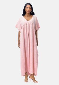 Robe longue rose texturée avec un col en V et des manches courtes. Forme fluide, fentes latérales jusqu'aux genoux, portée avec des talons rose clair.