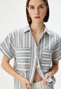 Camicia leggera a maniche corte con bottoni, a righe verticali di blu, rosa e bianco. Presenta due tasche e una texture morbida.