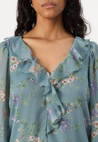 Blouse florale en tissu bleu clair, avec un décolleté en V à volants et des manches bouffantes. Des fleurs en orange, violet et vert créent des motifs délicats.
