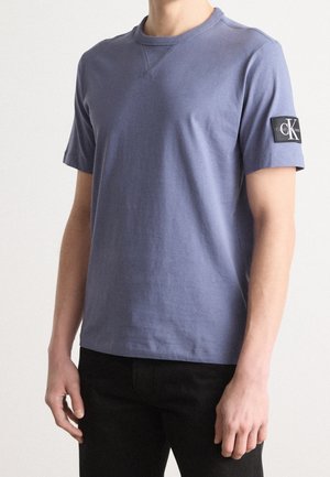 Person iført en simpel blå T-shirt med korte ærmer med et sort rektangulært logopatch på venstre ærme, kombineret med sorte bukser.