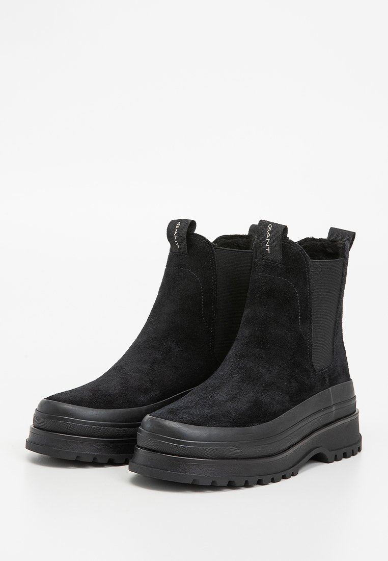 Botas de tobillo de ante negro con paneles elásticos a los lados, una suela gruesa negra y una lengüeta en la parte trasera para ponerlas y quitarlas fácilmente.