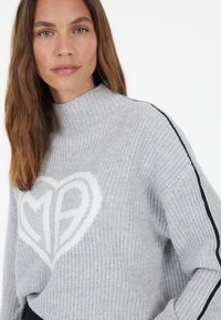 Grau gestrickter Pullover mit einem weißen Herz und dem Design "MP" in der Mitte sowie schwarzen Streifen entlang der Ärmel. Hoher Kragen.